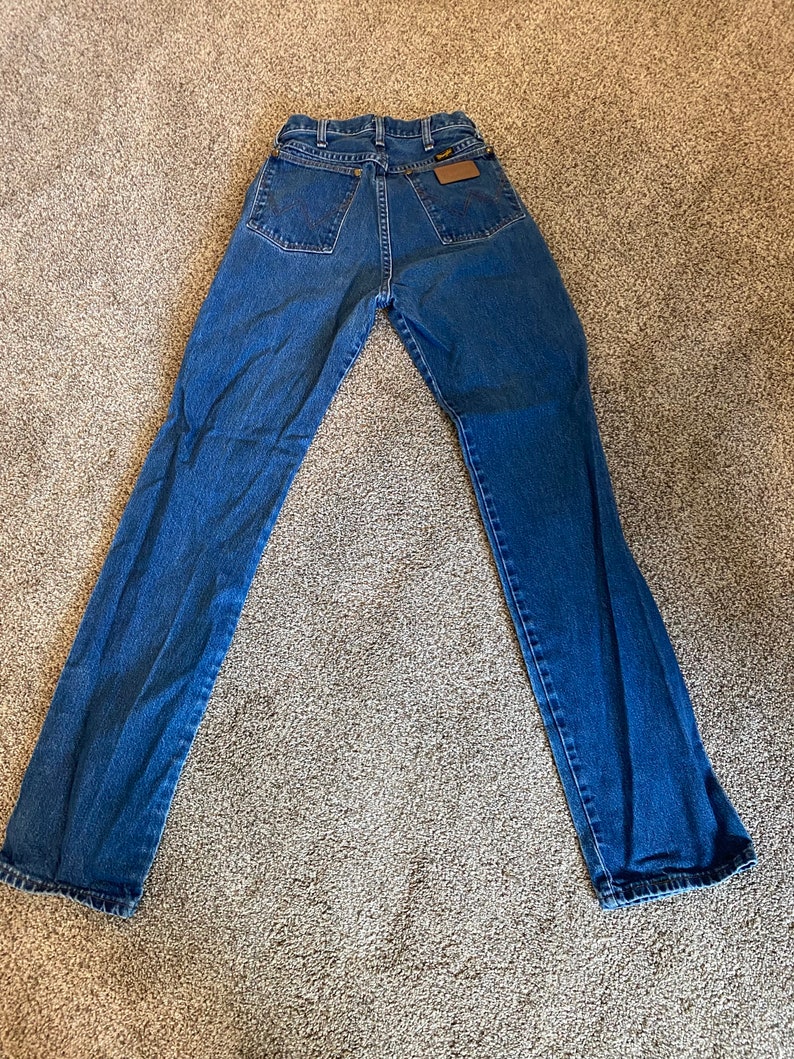 May include: Blue denim jeans with a classic five-pocket design. The jeans have a straight leg cut and a brown leather patch on the back. The jeans are laid flat on a neutral-colored surface.