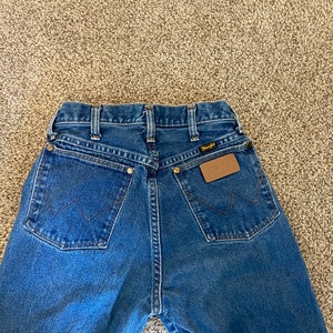 May include: Blue denim Wrangler jeans, viewed from the back. Features include classic five-pocket styling, a brown leather patch, and the Wrangler logo. The jeans have a straight leg cut and are made of durable denim.