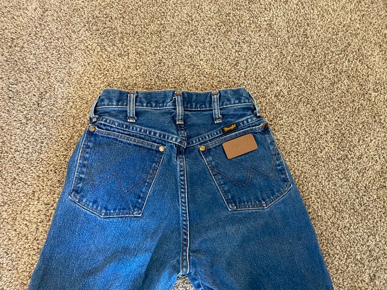 May include: Blue denim Wrangler jeans, viewed from the back. Features include classic five-pocket styling, a brown leather patch, and the Wrangler logo. The jeans have a straight leg cut and are made of durable denim.