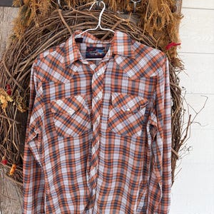 May include: A long-sleeved plaid shirt in shades of orange, navy, and white, hanging on a white hanger. The shirt features two chest pockets and a button-down front. The shirt is displayed against a rustic wooden background with a woven wreath.