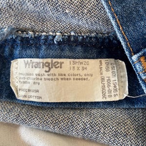 K&ouml;nnte beinhalten: Nahaufnahme eines blauen Jeanskleidungsst&uuml;cks mit einem wei&szlig;en Etikett. Das Etikett tr&auml;gt die Aufschrift "Wrangler" und enth&auml;lt Waschanweisungen und Gr&ouml;&szlig;eninformationen. Das Kleidungsst&uuml;ck besteht aus 100% Baumwolle.