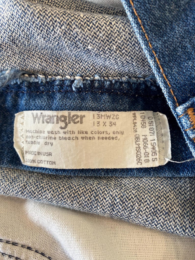 K&ouml;nnte beinhalten: Nahaufnahme eines blauen Jeanskleidungsst&uuml;cks mit einem wei&szlig;en Etikett. Das Etikett tr&auml;gt die Aufschrift "Wrangler" und enth&auml;lt Waschanweisungen und Gr&ouml;&szlig;eninformationen. Das Kleidungsst&uuml;ck besteht aus 100% Baumwolle.
