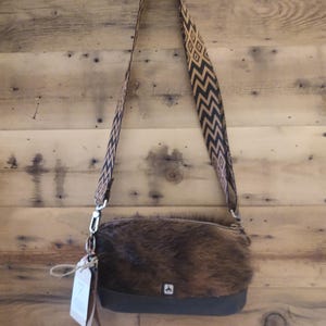 May include: A brown and black cowhide crossbody bag with a black and gold geometric patterned strap. The bag has a small flap with a metal buckle and a small tag attached to the strap.