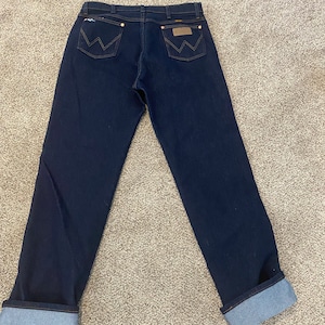 May include: A pair of dark blue denim jeans with a cuffed hem. The jeans have a brown leather patch with a white stitching design on the back.
