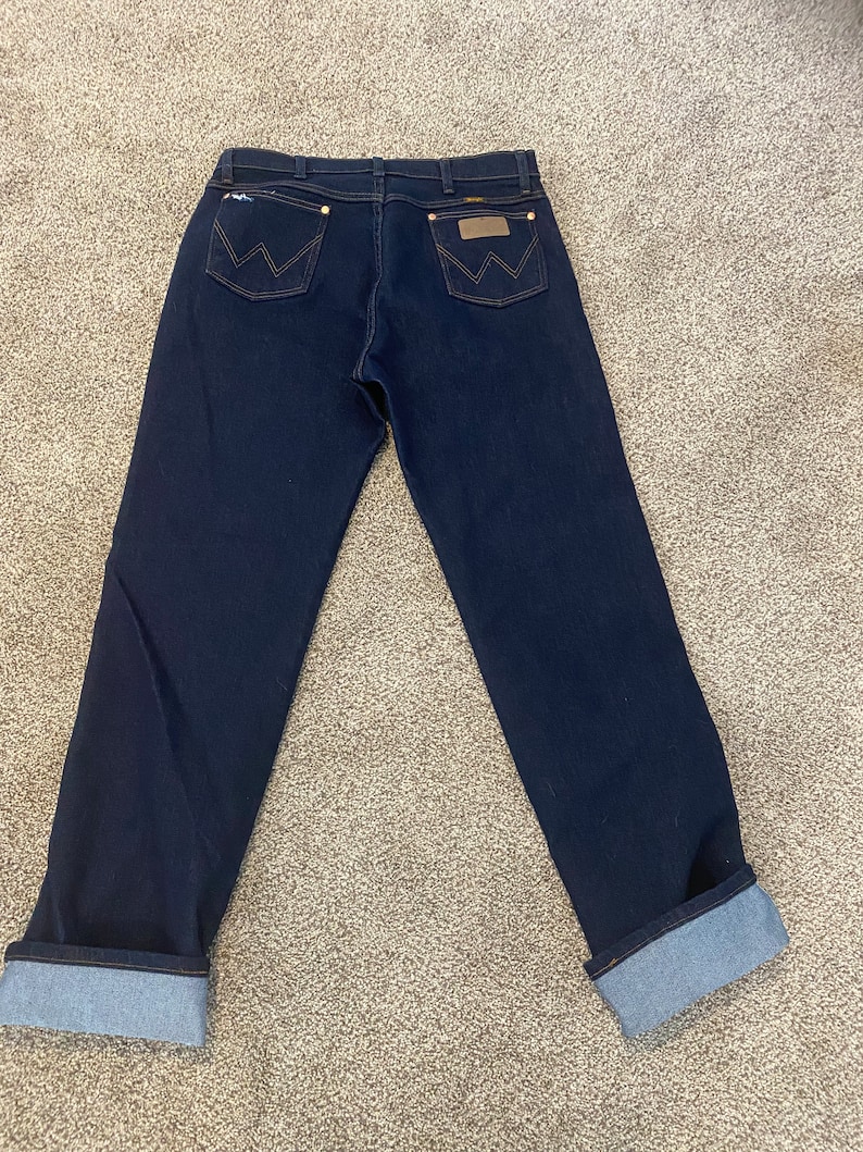 May include: A pair of dark blue denim jeans with a cuffed hem. The jeans have a brown leather patch with a white stitching design on the back.