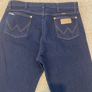 May include: A pair of dark blue denim jeans with a brown leather patch on the back pocket that says "Wrangler".