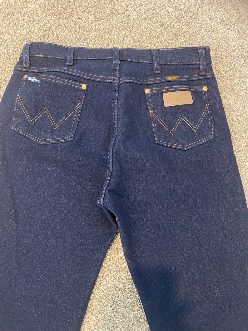 May include: A pair of dark blue denim jeans with a brown leather patch on the back pocket that says "Wrangler".