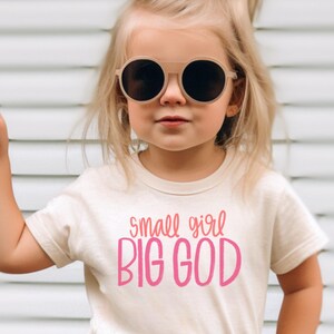 Puede incluir: Una ni&ntilde;a peque&ntilde;a con una camiseta de color crema con el texto "small girl BIG GOD" en letras rosas. Lleva gafas de sol redondas y tiene el pelo recogido en dos coletas con flores rosas.