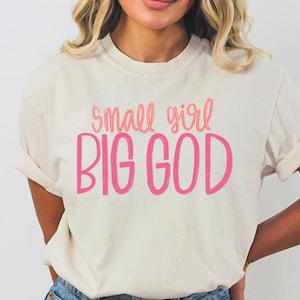 Puede incluir: Una camiseta blanca con el texto "small girl BIG GOD" en letras rosas.