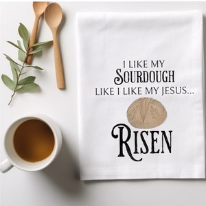May include: White kitchen towel with black text that reads "I like my sourdough like I like my Jesus... Risen". A brown sourdough bread loaf is pictured above the text.