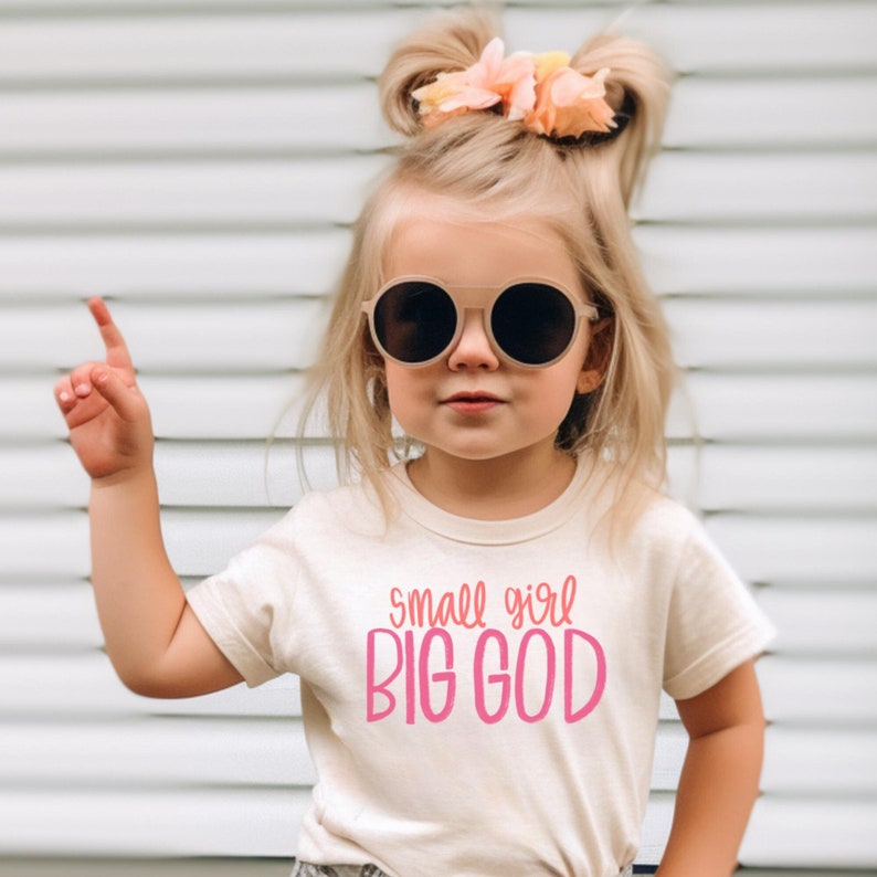 Puede incluir: Una ni&ntilde;a peque&ntilde;a con una camiseta de color crema con el texto "small girl BIG GOD" en letras rosas. Lleva gafas de sol redondas y tiene el pelo recogido en dos coletas con flores rosas.