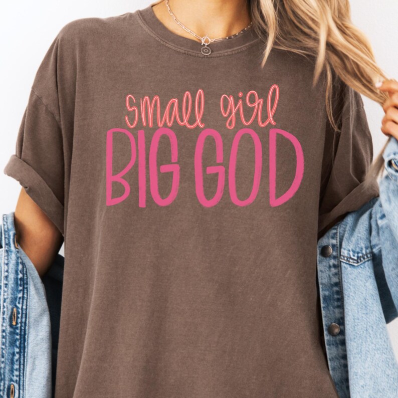 Puede incluir: Una camiseta marr&oacute;n con el texto "small girl BIG GOD" en letras rosas.
