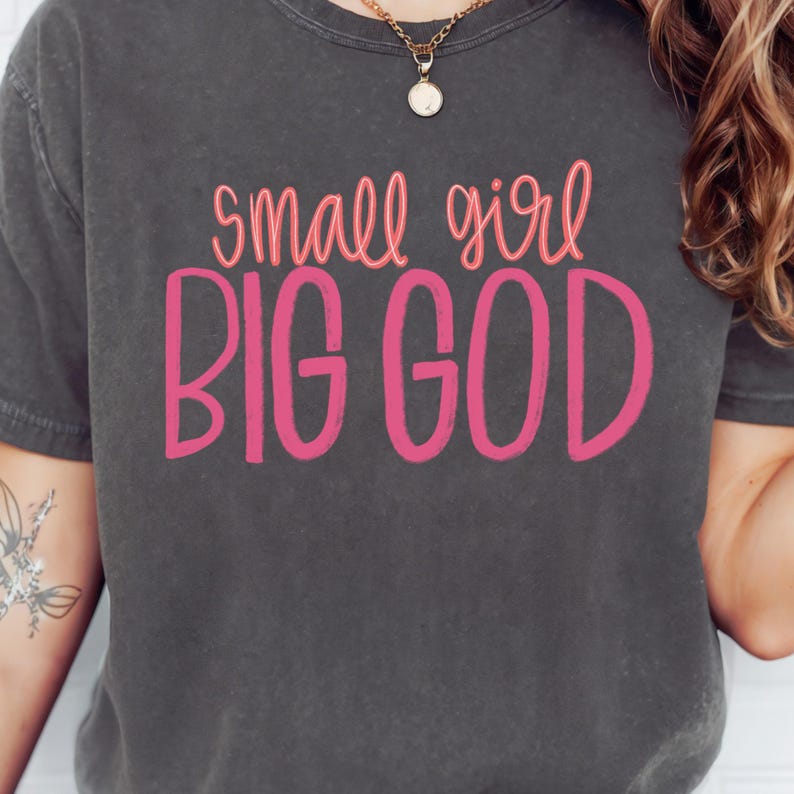 Puede incluir: Una camiseta gris oscura con el texto "small girl BIG GOD" en letras rosas.