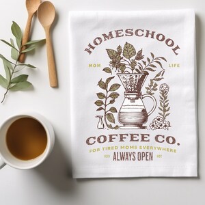 Puede incluir: Toalla de cocina blanca con una ilustraci&oacute;n marr&oacute;n de una cafetera con filtro y granos de caf&eacute;. El texto "HOMESCHOOL MOM LIFE" est&aacute; por encima de la ilustraci&oacute;n y "COFFEE CO. FOR TIRED MOMS EVERYWHERE" est&aacute; por debajo de la ilustraci&oacute;n. El texto "ICED ALWAYS OPEN HOT" est&aacute; debajo del nombre de la empresa de caf&eacute;.