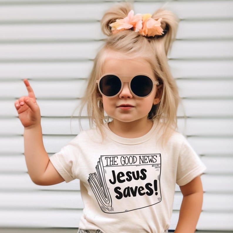 Pode incluir: Uma menina pequena vestindo uma camiseta creme com um gr&aacute;fico preto e branco de uma manchete de jornal que diz "The Good News Jesus Saves!" A menina est&aacute; usando &oacute;culos redondos e tem o cabelo puxado para tr&aacute;s com uma flor rosa.