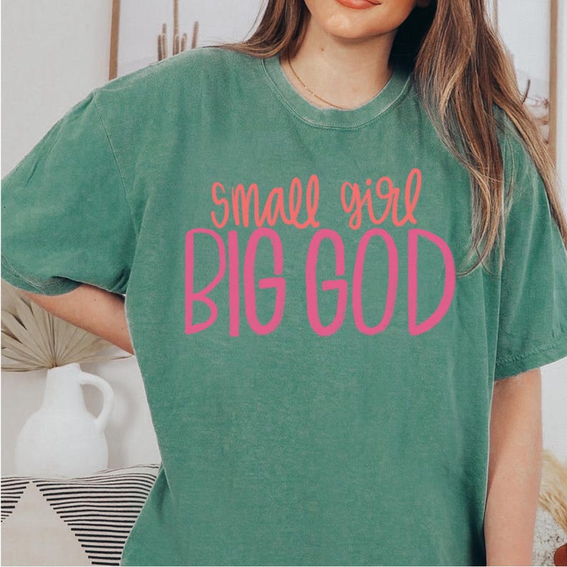 Puede incluir: Una camiseta verde con el texto "small girl BIG GOD" en letras rosas.