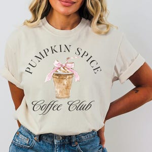 Könnte beinhalten: Ein weißes T-Shirt mit einem Aufdruck einer Kaffeetasse mit Schlagsahne und einer rosa Schleife. Der Text "Pumpkin Spice Coffee Club" ist auf das T-Shirt gedruckt.