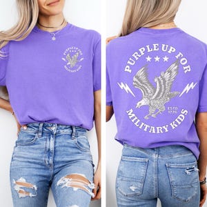 May include: A purple t-shirt with a graphic on the front and back. The front graphic says "PURPLE UP FOR MILITARY KIDS" with an eagle. The back graphic is the same, with the addition of "ESTD 1776".