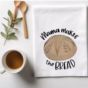 May include: White kitchen towel with the text "Mama makes the bread" in black cursive font. A brown loaf of bread with a cross pattern on top is illustrated in the center of the towel.
