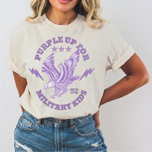 May include: Cream-colored t-shirt with a purple eagle graphic. The design includes the text "PURPLE UP FOR MILITARY KIDS" and "ESTD 1776". The eagle is depicted with outstretched wings, surrounded by stars and lightning bolts.