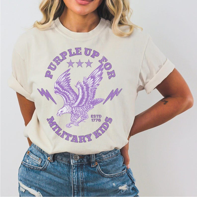 May include: Cream-colored t-shirt with a purple eagle graphic. The design includes the text "PURPLE UP FOR MILITARY KIDS" and "ESTD 1776". The eagle is depicted with outstretched wings, surrounded by stars and lightning bolts.