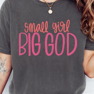 Puede incluir: Una camiseta gris oscura con el texto "small girl BIG GOD" impreso en rosa en la parte delantera.