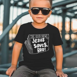 Pode incluir: Uma camiseta preta com um gr&aacute;fico branco de uma manchete de jornal que diz "The Good News: Jesus Saves, Bro!"