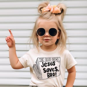 Pode incluir: Uma menina pequena vestindo uma camiseta bege com um gr&aacute;fico preto e branco que diz "The Good News Jesus Saves, Bro!" A menina est&aacute; usando &oacute;culos de sol redondos e tem o cabelo puxado para tr&aacute;s em duas tran&ccedil;as com uma flor rosa.