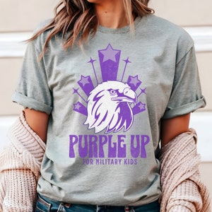 May include: Purple Up for Military Kids t-shirt featuring a purple eagle graphic and stars. The shirt is on a grey background.