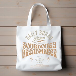 May include: White canvas tote bag with brown text that reads "Daily Bread Kitchen Baked with Love Sourdough Breadmaker Feed the Starter" with a lightning bolt graphic and wheat stalks.