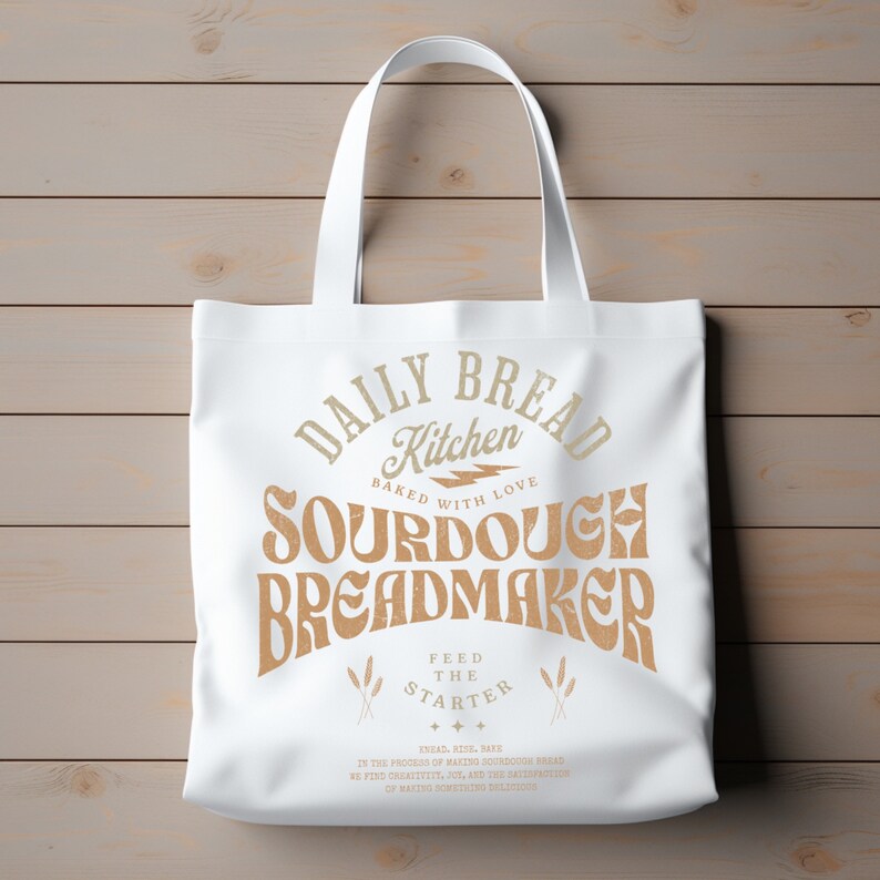 May include: White canvas tote bag with brown text that reads "Daily Bread Kitchen Baked with Love Sourdough Breadmaker Feed the Starter" with a lightning bolt graphic and wheat stalks.