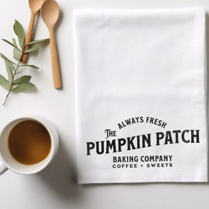 May include: White kitchen towel with black text that reads "The Always Fresh Pumpkin Patch Baking Company Coffee + Sweets".