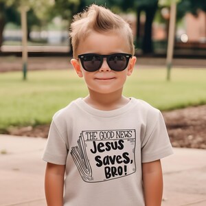 Pode incluir: Um menino pequeno vestindo uma camiseta bege com o texto "The Good News Jesus Saves, Bro!" impresso nela. Ele est&aacute; usando &oacute;culos de sol pretos e tem um penteado espetado.