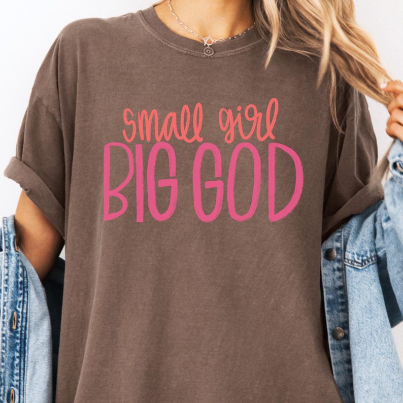 Puede incluir: Una camiseta marr&oacute;n con el texto "small girl BIG GOD" en letras rosa y naranja.
