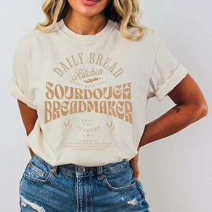 May include: A white t-shirt with a brown graphic that says "Daily Bread Kitchen Baked with Love Sourdough Breadmaker Feed the Starter" with a wheat design on the bottom.