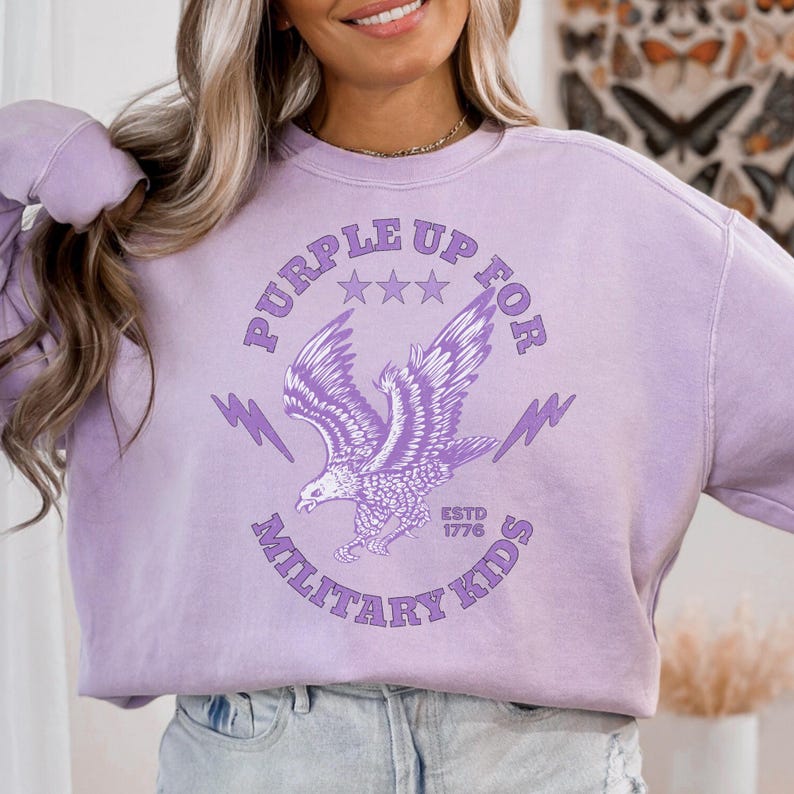 May include: A light purple sweatshirt with the text "PURPLE UP FOR MILITARY KIDS" and an eagle graphic. The sweatshirt has a crew neck and long sleeves. The text "ESTD 1776" is also visible.