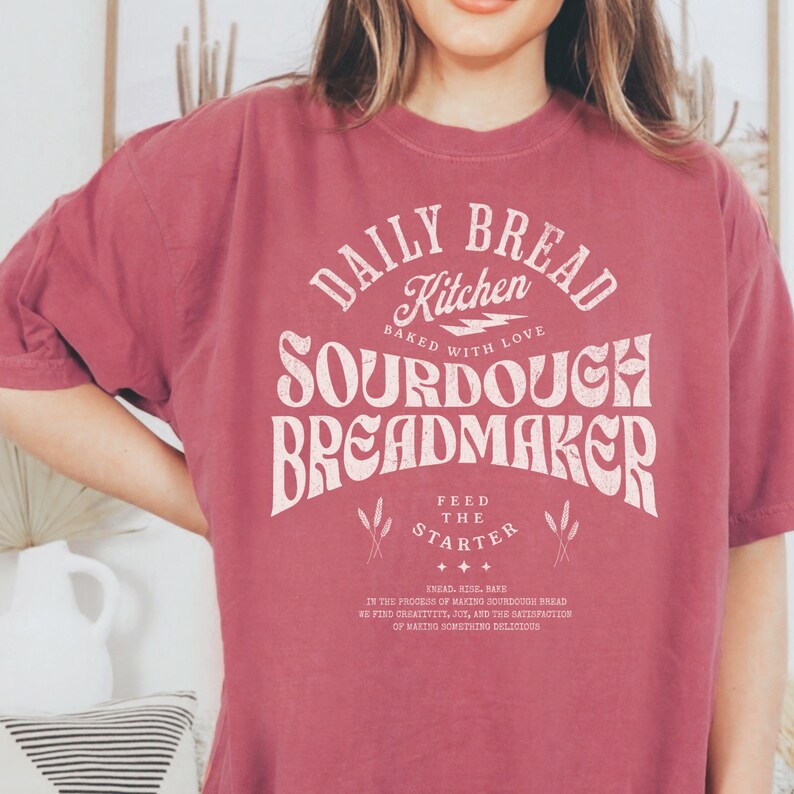 May include: A maroon t-shirt with a white graphic design that says "Daily Bread Kitchen Baked with Love Sourdough Breadmaker Feed the Starter Knead, Rise, Bake In the process of making sourdough bread we find creativity, joy, and the satisfaction of making something delicious."