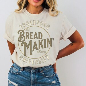 May include: A white t-shirt with a brown circle graphic that says "The Bread Makin' Mama University" in a vintage font.