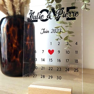 Könnte beinhalten: Transparenter Acrylkalender mit den Namen "Julie & Pierre" und dem Wort "LOVE" in schwarzer Schrift. Der Kalender zeigt den Monat Juni 2023, mit einem roten Herz am 8. Er steht auf einem Holzsockel.
