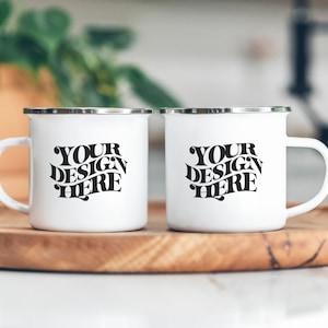 May include: Two white enamel mugs with silver rims. The mugs have black text that reads "YOUR DESIGN HERE" on each mug.