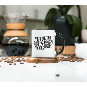 May include: White ceramic coffee mug with a black rim and handle. The mug has the text "YOUR DESIGN HERE" printed in black on the front. The mug is sitting on a wooden coaster and there are coffee beans scattered around it.