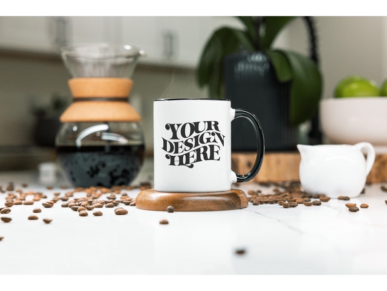 May include: White ceramic coffee mug with a black rim and handle. The mug has the text "YOUR DESIGN HERE" printed in black on the front. The mug is sitting on a wooden coaster and there are coffee beans scattered around it.