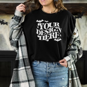Puede incluir: Una camiseta negra con un gráfico blanco que dice "YOUR DESIGN HERE" con murciélagos y estrellas alrededor del texto. La camiseta la lleva puesta una persona con una camisa de cuadros encima.
