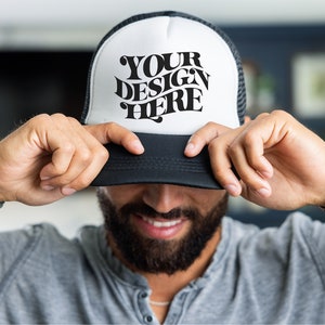May include: A black and white trucker hat with a white front panel and a black mesh back. The front panel has the text "YOUR DESIGN HERE" in a black retro font.