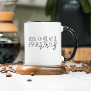 May include: White ceramic mug with a black rim and handle. The mug has the text "model mockup company" printed on it in gray. The mug is sitting on a wooden coaster.