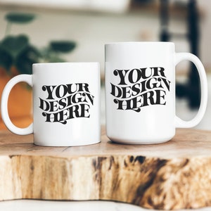 Puede incluir: Dos tazas de cerámica blanca con texto negro que dice "YOUR DESIGN HERE" sobre una superficie de madera.