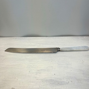 Puede incluir: Un cuchillo de pan largo y serrado con una hoja de metal plateado que muestra un logotipo grabado y un mango blanco nacarado liso. El cuchillo presenta un refuerzo distintivo que conecta la hoja con el mango, creando un diseño elegante.