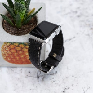 以下が含まれることがあります： 白いステッチが施された黒い革バンドが付いたシルバーのApple Watch。パイナップルのデザインと多肉植物が施されたコンクリートプランターの隣に時計が表示されています。