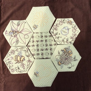 May include: A green and white hexagon-shaped quilt block with an embroidered bee theme. The block features bees, honeycombs, flowers, and the word "Honey".