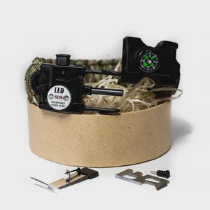Könnte beinhalten: Ein Survival-Kit mit einem schwarzen Kompass, LED SOS-Licht und einem Tarnfarben-Paracord-Armband. Das Kit enthält ein kleines Messer, ein Multi-Tool und ein kleines schwarzes Teil. Das Kit befindet sich in einem runden Pappbehälter.